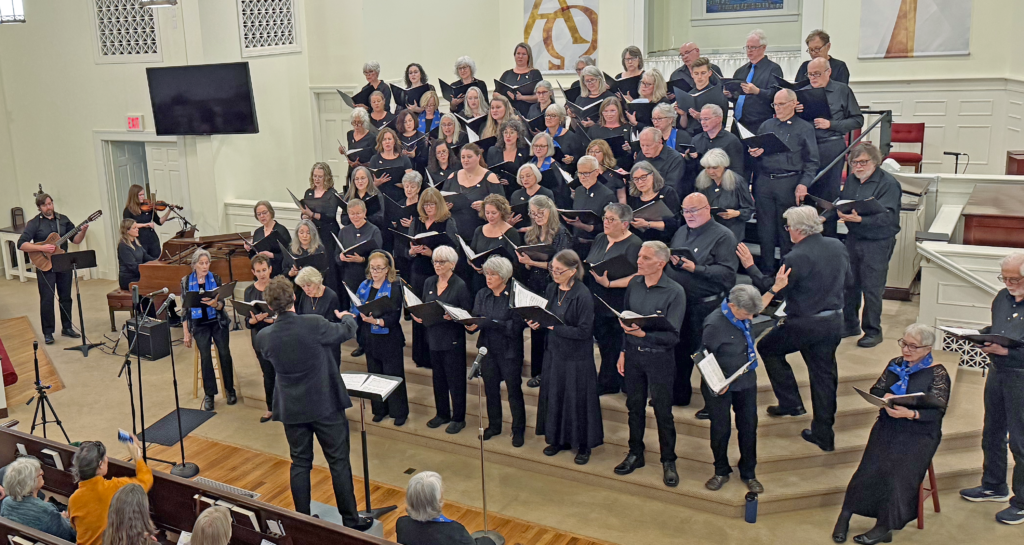 Crozet Community Chorus performing at their April 2026 concert in Crozet, Virginia. Choristers dressed in black stand on risers in a church, accompanied by piano, guitar, and violin